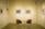 A minimalist art gallery setting features a small round table with two curved wooden chairs. The walls display several black and white framed photographs. The room is illuminated by track lighting, creating a serene and contemplative atmosphere.