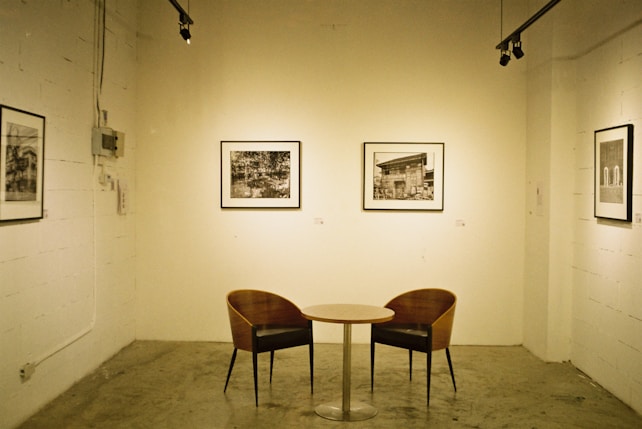 A minimalist art gallery setting features a small round table with two curved wooden chairs. The walls display several black and white framed photographs. The room is illuminated by track lighting, creating a serene and contemplative atmosphere.