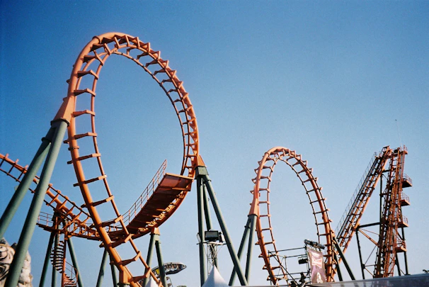 A vibrant amusement park roller coaster featuring multiple loops rises against a clear blue sky. The structure is orange and supported by sturdy green beams. The intricate design and engineering are highlighted by the sun's light.