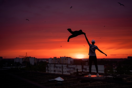 man in blue t-shirt and black shorts standing on roof top during sunset