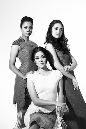 Three elegantly dressed women pose in a studio setting. The image is in black and white, highlighting the sophisticated and formal attire they are wearing, along with their poised expressions and hairstyles.