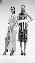 Two women stand confidently in intricate, patterned dresses and high heels. One dress features wrap elements with a high slit, while the other is adorned with a large white belt. The setting is minimal, enhancing the focus on their fashion and stance.