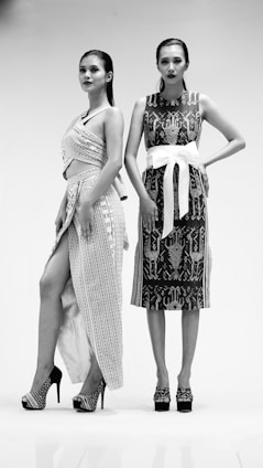 Two women stand confidently in intricate, patterned dresses and high heels. One dress features wrap elements with a high slit, while the other is adorned with a large white belt. The setting is minimal, enhancing the focus on their fashion and stance.