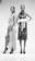 Two women stand confidently in intricate, patterned dresses and high heels. One dress features wrap elements with a high slit, while the other is adorned with a large white belt. The setting is minimal, enhancing the focus on their fashion and stance.