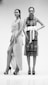 Two women stand confidently in intricate, patterned dresses and high heels. One dress features wrap elements with a high slit, while the other is adorned with a large white belt. The setting is minimal, enhancing the focus on their fashion and stance.