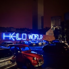 A nighttime urban scene with a prominent neon blue 'HELLO WORLD' sign on a brick wall. Several parked cars, including a red vintage car, are visible in the foreground under dim street lighting. The silhouettes of people standing near the cars add a mysterious and modern ambiance.