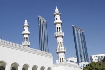 A mosque with intricate minarets is seen in the foreground, characterized by white and ornate architectural details. Modern skyscrapers with reflective glass surfaces stand in the background under a clear blue sky.