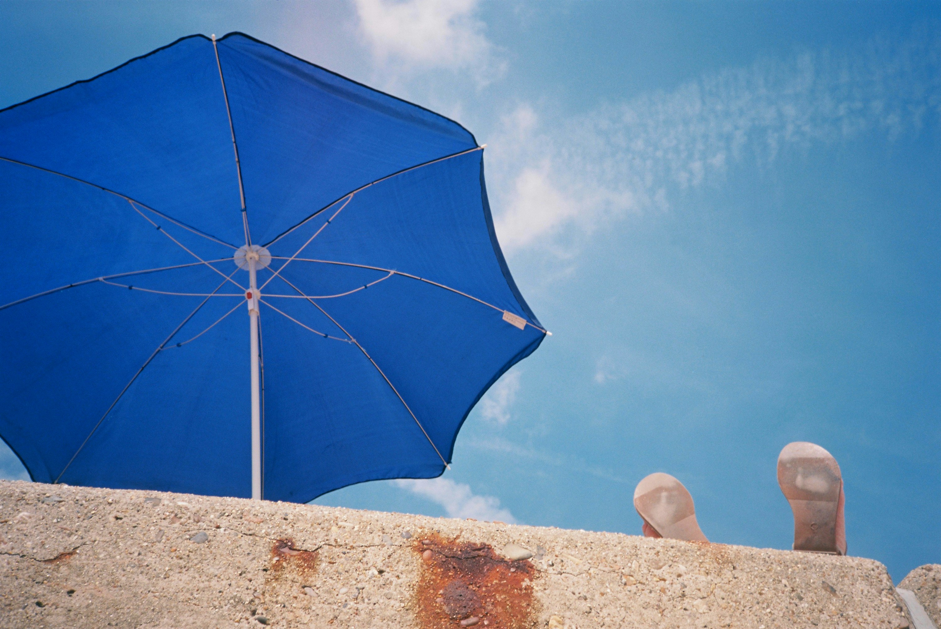 blue umbrella on white sand, A person relaxing by putting their feet up on a sea wall. They are beside a blue parasol or beach umbrella at the seaside under a blue summer sky. Shot on film.