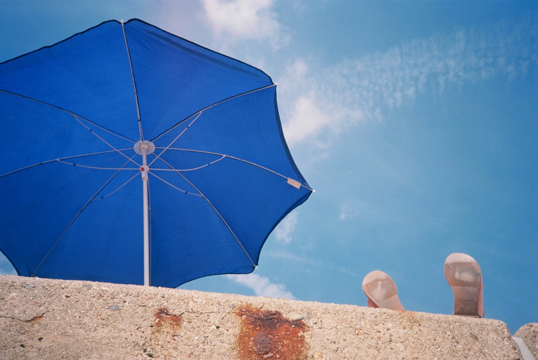 blue umbrella on white sand, A person relaxing by putting their feet up on a sea wall. They are beside a blue parasol or