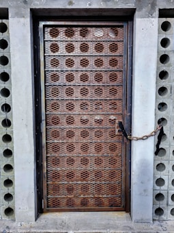 A metal door with a rusted grid pattern is secured with a chain and lock against a concrete wall. The surrounding wall has a series of symmetrical circular cutouts on either side of the door.