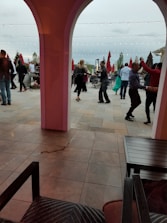 People are dancing in an outdoor patio area, surrounded by tables and large red umbrellas. Strings of lights are hung above, creating a festive ambiance. The setting appears to be lively and social.
