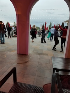 People are dancing in an outdoor patio area, surrounded by tables and large red umbrellas. Strings of lights are hung above, creating a festive ambiance. The setting appears to be lively and social.
