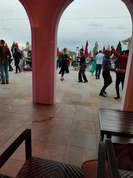People are dancing in an outdoor patio area, surrounded by tables and large red umbrellas. Strings of lights are hung above, creating a festive ambiance. The setting appears to be lively and social.