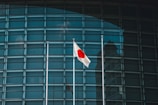 A Japanese flag is prominently displayed, fluttering on a flagpole in front of a modern building with a grid-like glass facade. The reflection of a nearby structure is visible in the glass panels.