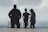 Three individuals are standing on a platform overlooking a vast, cloudy landscape. The figures appear as silhouettes against the bright sky, with letters spelling 'PUK' visible in the background.