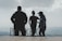 Three individuals are standing on a platform overlooking a vast, cloudy landscape. The figures appear as silhouettes against the bright sky, with letters spelling 'PUK' visible in the background.