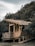 A rustic wooden cabin situated against a dense backdrop of trees, with a weathered exterior and simple architecture. The cabin features an elevated platform with wooden railings and a slanted roof.