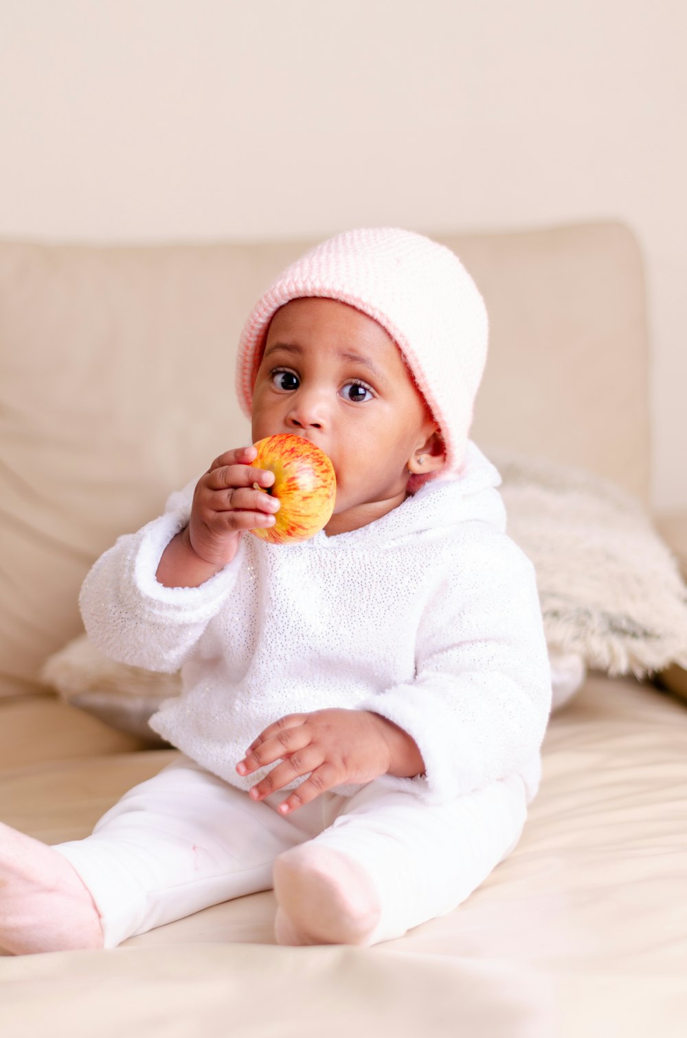 baby in white knit cap holding orange fruit
