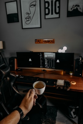 A cozy workspace with a wooden desk, dual monitors, and a comfortable leather chair. Above the desk are three pieces of wall art, including an abstract line drawing of a face and the word 'BIG'. A hand holding a small espresso cup is positioned in the foreground, adding a personal touch. The desk is neatly organized with a lamp, a plant, and various desk accessories.