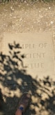 A stone plaque on the ground with the engraving 'TEMPLE OF ANCIENT VIRTUE'. Shadows of leaves cast patterns over the plaque, and a brown shoe is partially visible at the bottom.