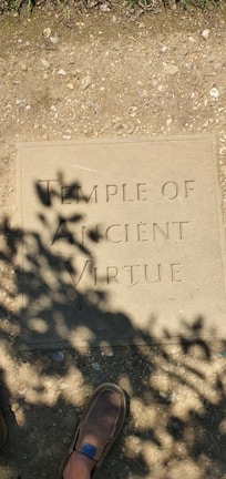 A stone plaque on the ground with the engraving 'TEMPLE OF ANCIENT VIRTUE'. Shadows of leaves cast patterns over the plaque, and a brown shoe is partially visible at the bottom.