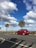 A bright red car is driving along a coastal road lined with palm trees. The sky is predominantly blue with large, fluffy white clouds scattered throughout. Shadows of palm fronds are cast on the road.