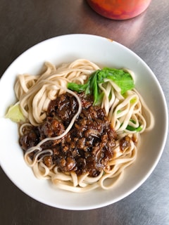A white bowl filled with thick noodles topped with a savory, dark brown sauce and green leafy vegetables, placed on a metallic table surface. The noodles are neatly arranged, showcasing the glossy texture of the sauce. A hint of a red container partially appears in the background.