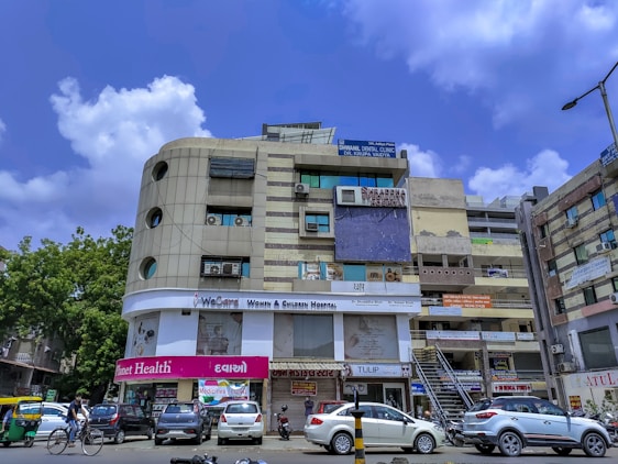 A multi-story building with various business signs and advertisements, including healthcare-related establishments like dental and surgical clinics. The structure has a modern facade with windows and air conditioning units. Parked cars and a motorcycle, as well as a cyclist, are visible on the street. The sky is clear with a few clouds.