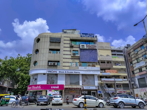 A multi-story building with various business signs and advertisements, including healthcare-related establishments like dental and surgical clinics. The structure has a modern facade with windows and air conditioning units. Parked cars and a motorcycle, as well as a cyclist, are visible on the street. The sky is clear with a few clouds.