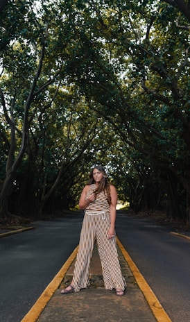 woman in brown dress standing on road between trees during daytime