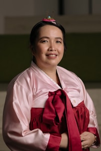 A person is wearing a traditional Korean hanbok, featuring a light pink jeogori with a deep red ribbon and skirt. The hair is styled with an ornamental hairpiece that includes a small, colorful embellishment. The person is smiling gently, conveying a calm and pleasant demeanor.
