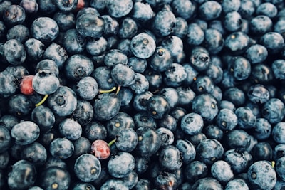 A close-up view of a pile of fresh blueberries with a few red berries interspersed. The fruits are plump and exhibit a range of blue hues, some with slight frost on their surface.