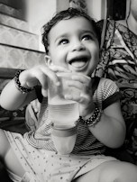 A smiling baby holding a clear bottle with measurement markings, sitting in a high chair.