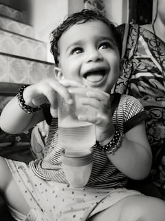 A smiling baby holding a clear bottle with measurement markings, sitting in a high chair.