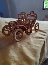 A handcrafted model of a vintage car made from woven straw sits on a beige fabric-covered table. The car model features intricate detailing with a lattice design on the seat and body, along with round wheels and bent straw for the handlebars.