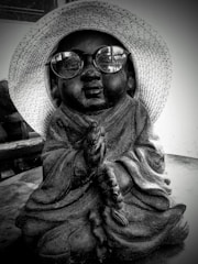 A statue of a child monk wearing a straw hat and round glasses is placed on a table. The monk is in a meditative posture, holding prayer beads. The image has a black and white filter, giving it a nostalgic and serene feel.