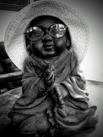 A statue of a child monk wearing a straw hat and round glasses is placed on a table. The monk is in a meditative posture, holding prayer beads. The image has a black and white filter, giving it a nostalgic and serene feel.