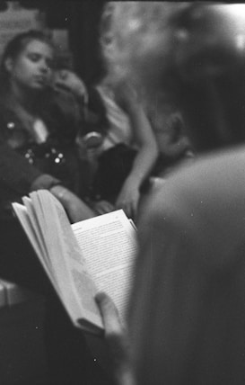 A black and white photograph features a close-up of a person holding an open book, with another person sitting in the background, slightly out of focus. The setting appears to be casual and possibly in a public place such as a caf&eacute; or public transport.