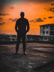 A dramatic still showing a lone figure silhouetted against a vibrant sunset on a sprawling urban rooftop.