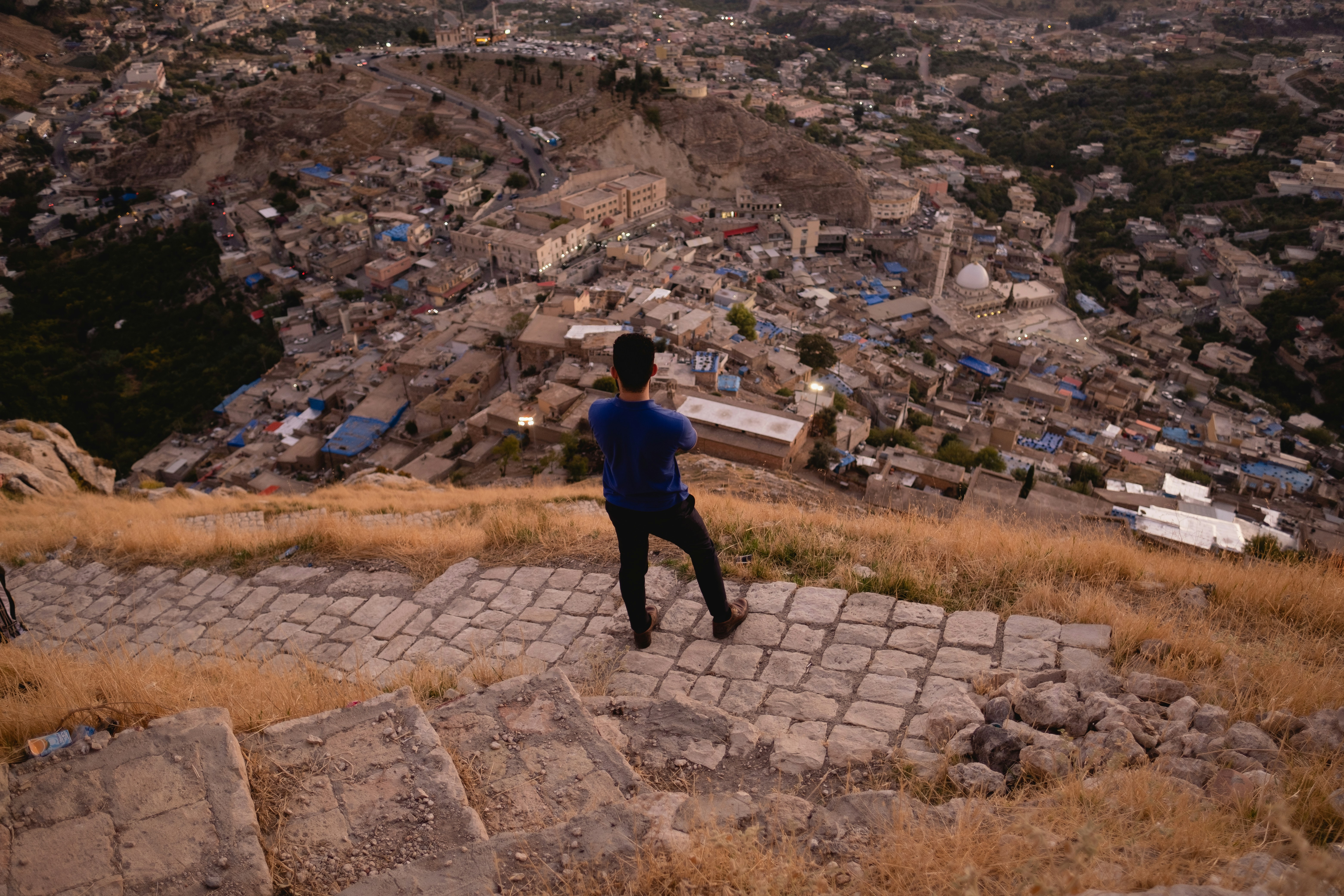 man in blue jacket standing on gray concrete brick floor during daytime, Overlooking the town of Akre, in the Kurdistan Region.