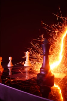 A dramatic scene on a chessboard with pieces including a black king at the forefront, surrounded by fiery orange and yellow sparks against a dark background. The light creates an intense, dynamic effect with reflections on the glossy surface.