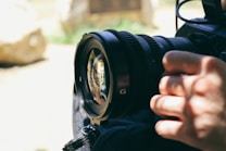 A close-up of a camera lens with a person's hand adjusting it. The background is softly blurred, suggesting an outdoor setting with natural light. The lens reflects some of the surroundings, showcasing the precision and detail of the equipment.