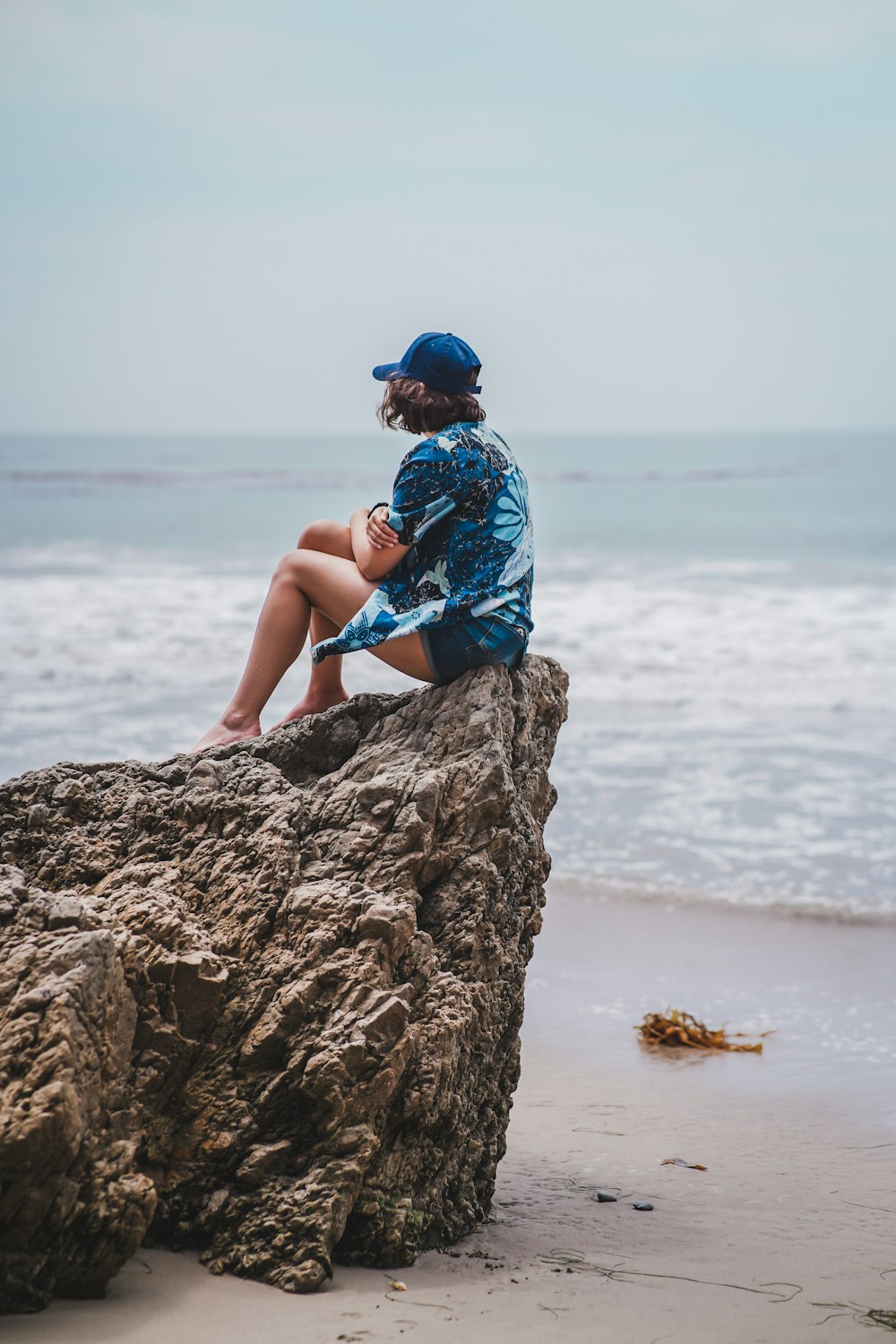 femme en robe fleurie bleue et blanche assise sur le rocher près de la mer pendant la journée