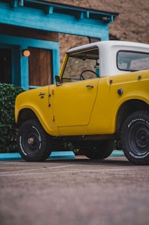 A vintage yellow off-road vehicle with a white roof is parked outside a light blue building with a wooden door. The vehicle has large tires and appears to be in good condition. There is greenery next to the building, and the ground appears to be paved.
