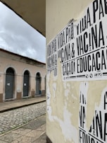 A street corner featuring a wall covered with black and white posters that have messages in Portuguese. The building in the background has pale pink walls and several closed shutters, indicating it may be a shop or residential area. The street is paved with cobblestones and appears deserted under a cloudy sky.