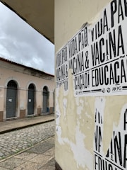 A street corner featuring a wall covered with black and white posters that have messages in Portuguese. The building in the background has pale pink walls and several closed shutters, indicating it may be a shop or residential area. The street is paved with cobblestones and appears deserted under a cloudy sky.