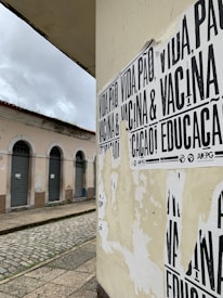 A street corner featuring a wall covered with black and white posters that have messages in Portuguese. The building in the background has pale pink walls and several closed shutters, indicating it may be a shop or residential area. The street is paved with cobblestones and appears deserted under a cloudy sky.