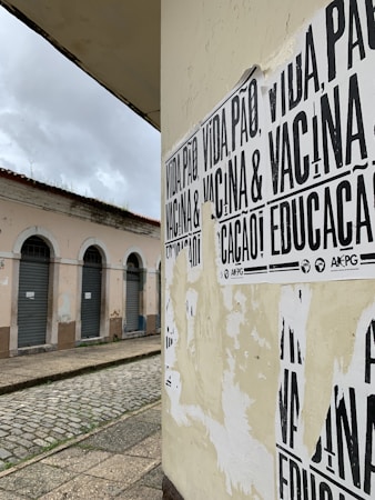 A street corner featuring a wall covered with black and white posters that have messages in Portuguese. The building in the background has pale pink walls and several closed shutters, indicating it may be a shop or residential area. The street is paved with cobblestones and appears deserted under a cloudy sky.
