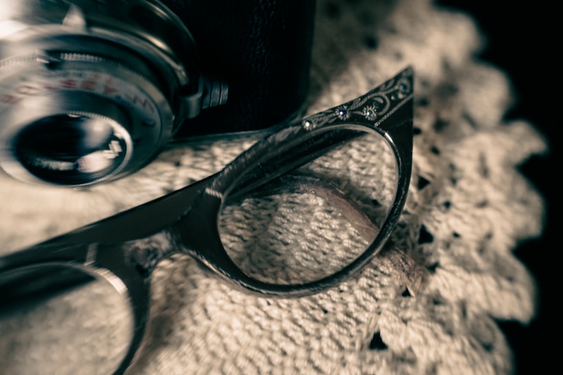 A close-up shot of a vintage camera lens and a pair of retro glasses with intricately designed frames. The items are placed on a textured, white crocheted fabric, creating an antique aesthetic.