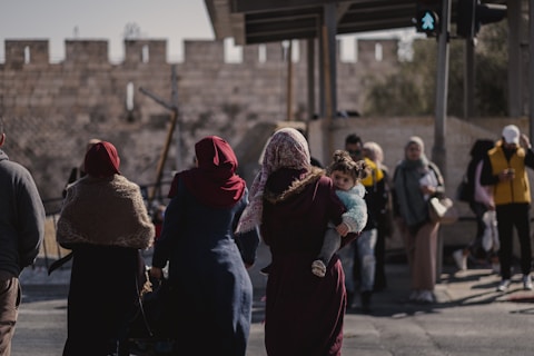 A group of people are walking in an urban setting near an old stone wall. One person is carrying a child, and others are dressed in warm clothing, suggesting cooler weather. The scene includes a variety of people, indicating a diverse community.